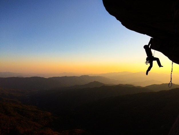 Vista de ángulo bajo de un hombre en silueta escalando una montaña rocosa durante la puesta de sol