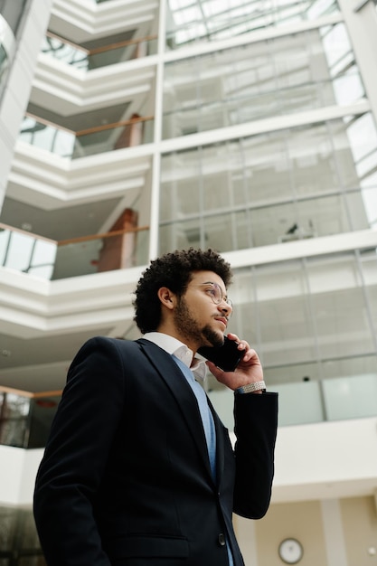 Vista de ángulo bajo del joven hombre de negocios en traje hablando por teléfono móvil mientras camina por la oficina h