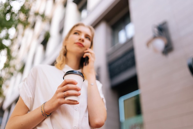 Vista de ángulo bajo de una joven mujer bonita con ropa informal hablando por teléfono inteligente mientras está de pie en la calle de la ciudad el día de verano