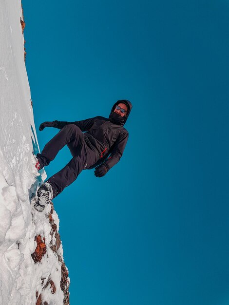 Foto vista de ángulo bajo de un joven de pie en una montaña cubierta de nieve contra un cielo azul claro