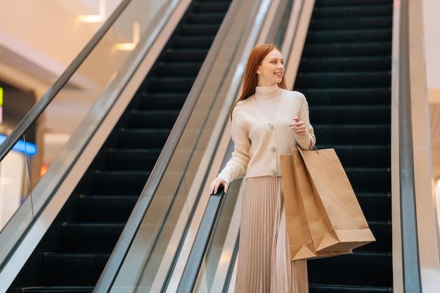 Vista de ángulo bajo de una joven sonriente sosteniendo el pasamanos de la escalera mecánica y montando la escalera mecánica subiendo en el centro comercial, mirando hacia otro lado, bolsas de papel con compras en las manos, fondo borroso.