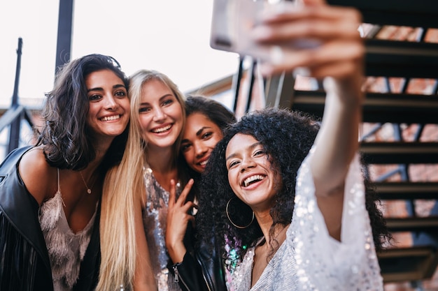 Vista de ángulo bajo de jóvenes amigas alegres tomando selfies con el teléfono móvil en la escalera