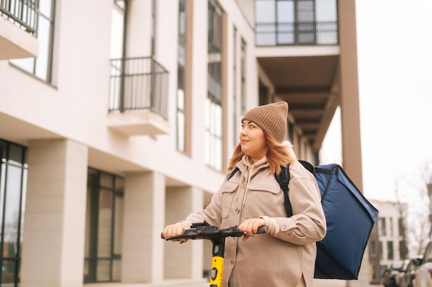 Vista de ángulo bajo de mensajero femenino con gran mochila térmica montando en scooter eléctrico en la calle de la ciudad mirando hacia otro lado