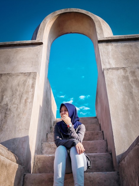 Foto vista de ángulo bajo de una mujer con hijab sentada en escalones contra el cielo azul