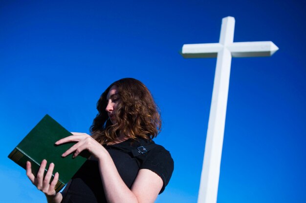 Foto vista de ángulo bajo de una mujer leyendo la biblia mientras está de pie en la cruz