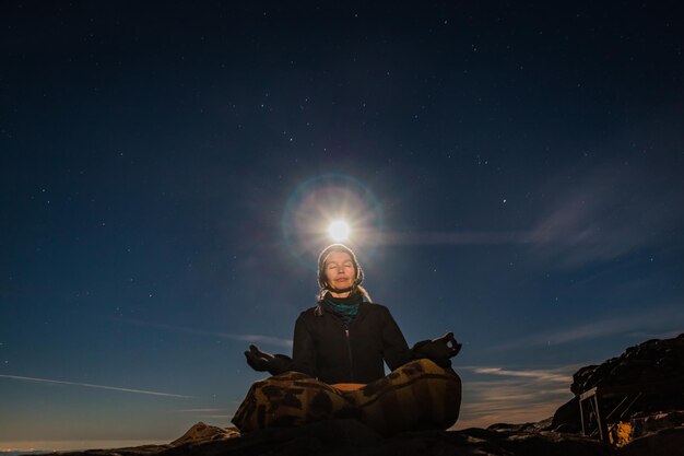 Foto vista de ángulo bajo de una mujer meditando mientras está sentada en una roca contra el cielo durante la puesta de sol