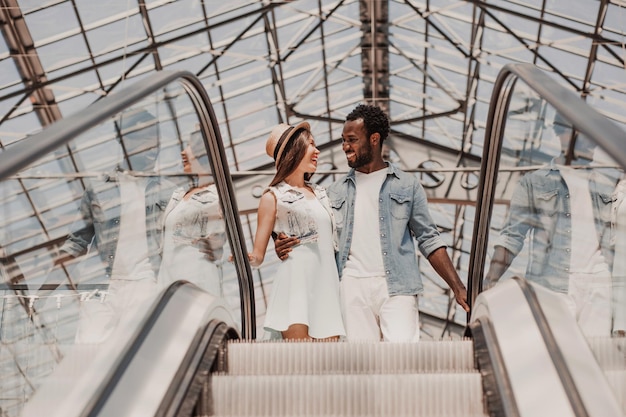 Vista de ángulo bajo de una pareja multiétnica sonriendo unos a otros de pie en la escalera mecánica en el centro comercial