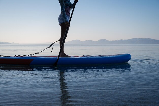 Vista de ángulo bajo de perfil de una mujer joven de pie en el tablero de sup remando en el mar tranquilo de la mañana