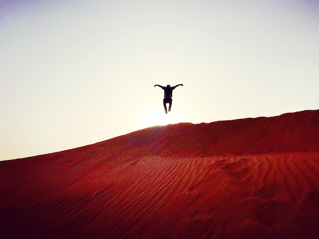 Foto vista de bajo ángulo de una persona con los brazos extendidos saltando sobre la arena en el desierto contra un cielo despejado
