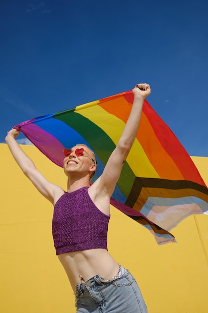 Vista de ángulo bajo de una persona no binaria feliz joven sosteniendo y agitando una bandera del arco iris sobre la cabeza. Concepto de igualdad, derechos e identidad de género.