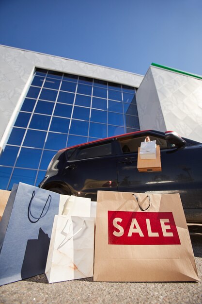 Vista de ángulo bajo vertical en bolsas de la compra con cartel de VENTA al aire libre contra el edificio del centro comercial y el coche en la superficie, espacio de copia