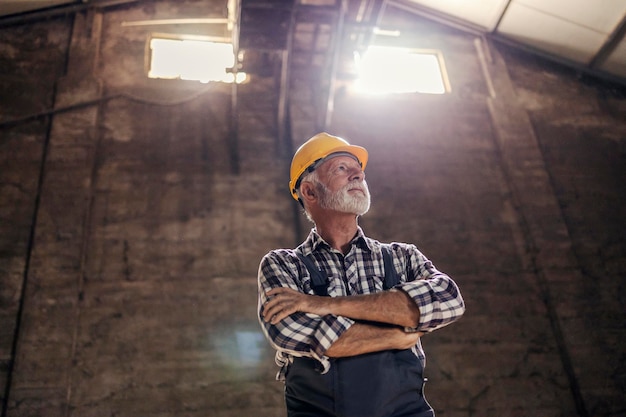 Foto vista de ángulo bajo de un viejo trabajador de barba blanca con las manos cruzadas esperando la próxima tarea en la fábrica