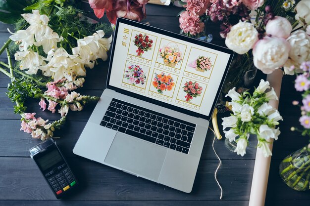 Vista anterior de la computadora portátil abierta con el sitio web de la florería en la pantalla, hermosas flores y terminal de pago en la mesa en la florería