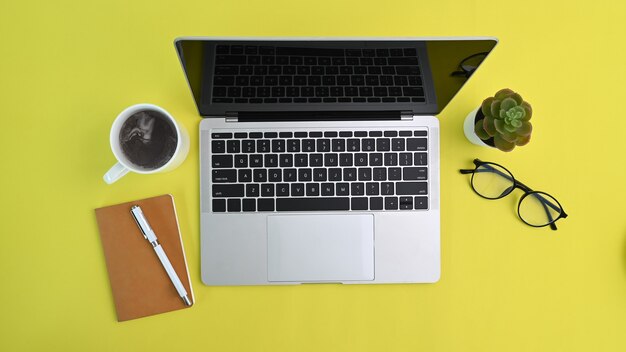 Vista anterior de la computadora portátil, taza de café, vasos y cuaderno sobre fondo amarillo.