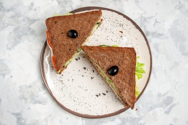 Vista anterior del delicioso sándwich con pan negro decorado con oliva en un plato sobre la superficie blanca manchada