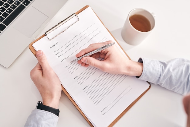 Vista anterior del gerente irreconocible sentado a la mesa con una taza y una computadora portátil y llenando la información personal en la tarjeta médica