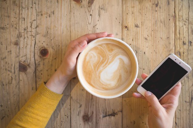 Vista anterior de manos femeninas sosteniendo una taza de café caliente y con un teléfono inteligente en la mesa de madera