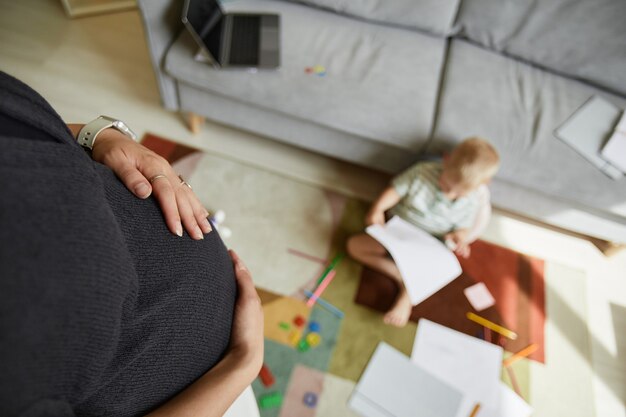 Vista anterior de una mujer embarazada irreconocible acariciando el vientre mientras su hijo está sentado en la alfombra y dibujando
