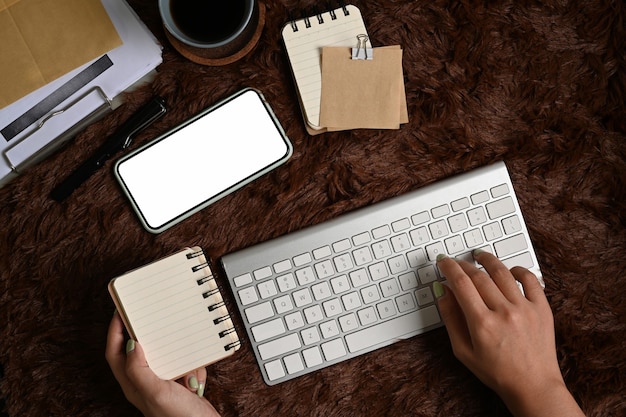 Vista anterior de una mujer escribiendo en un teclado inalámbrico y sosteniendo un cuaderno sobre una alfombra de piel marrón.