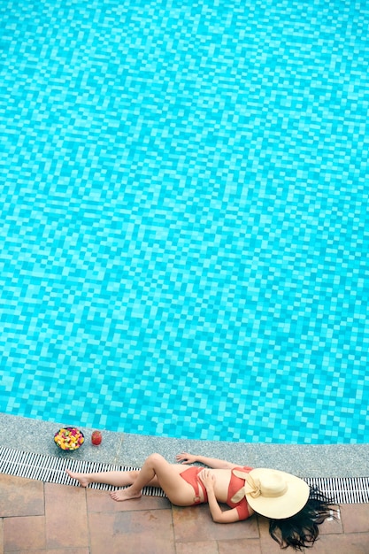 Foto vista anterior de una mujer irreconocible con sombrero de paja en la cara que yacía cerca de la piscina al aire libre