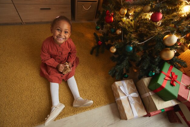 Vista anterior retrato de niña afroamericana emocionada abriendo regalos de Navidad mientras está sentado junto al árbol en casa y sonriendo a la cámara, espacio de copia