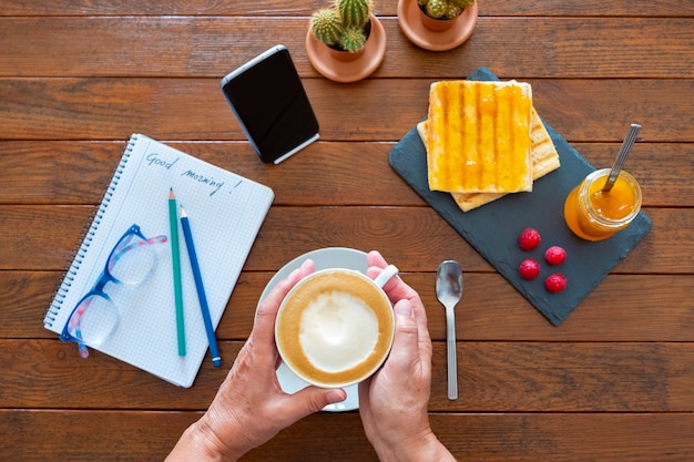 Vista anterior del rincón del desayuno con dos manos humanas sosteniendo una taza de capuchino con espuma de leche y rebanadas de pan y mermelada en una mesa de madera - Bloc de notas y vasos