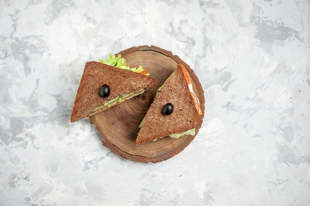 Vista anterior de sabroso sándwich con pan negro decorado con oliva sobre una tabla de cortar de madera sobre una superficie blanca manchada con espacio libre