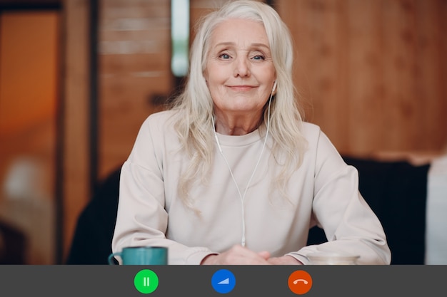 Vista de la aplicación en pantalla de una anciana sonriente sentada a la mesa con una computadora portátil y hablando por videollamada con un amigo o compañero de trabajo, una mujer madura feliz con una taza de té o café hablando en línea por una cámara web en el interior