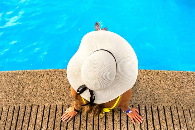 Vista de arriba hacia abajo de una mujer joven con sombrero de paja amarillo descansando cerca de la piscina con agua azul clara