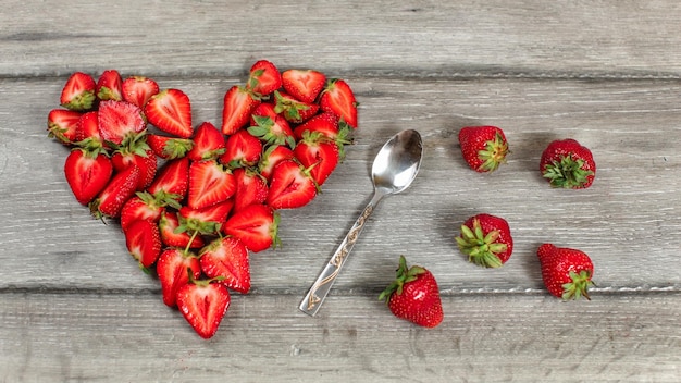 Vista de arriba hacia abajo, trozos de fresa dispuestos en forma de corazón, cuchara y algunas fresas derramadas cerca, sobre un escritorio de madera gris.