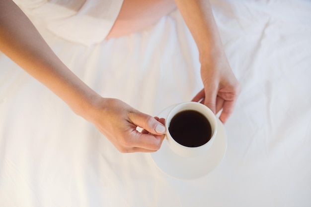 Vista desde arriba hermosa mujer relajándose en un acogedor ambiente hogareño en la cama blanca sosteniendo una taza de chocolate caliente o café en sus manos disfrutando de la comodidad Fondo de luz suave Vista superior