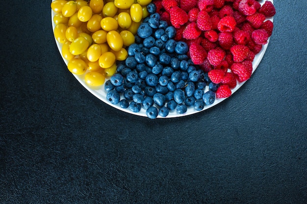 Vista desde arriba. Una mezcla de bayas amarillas, rojas y azules en un rallador blanco. Triángulo de separación. Mezcla de frutas de verano. Diseño de bayas