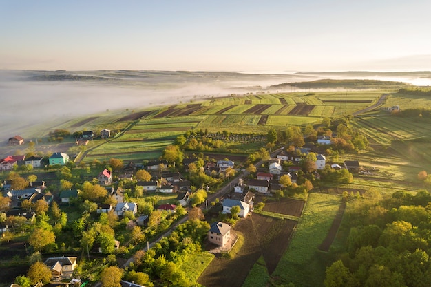 Vista desde arriba de la niebla blanca en los tejados de las casas de pueblo entre árboles verdes bajo un cielo azul brillante. Panorama de paisaje brumoso de primavera al amanecer.