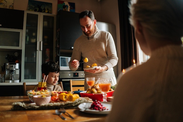 Vista desde atrás al joven que organiza la comida en platos para la esposa y el hijo de pie en la mesa festiva de Navidad durante la fiesta familiar