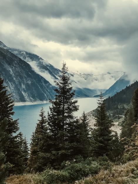 Vista através dos pinheiros para o lago de montanha turquesa claro Schlegays coberto de nuvens e nevoeiro. Alpes Zillertal, Mayrhofen, Áustria.