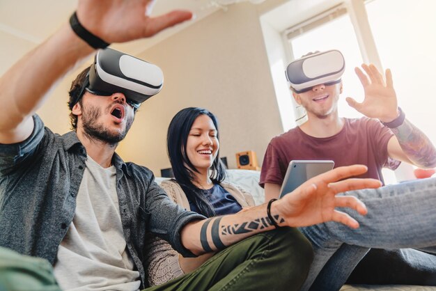 Foto vista baja de amigos sorprendidos divirtiéndose con gafas de realidad virtual en casa mientras están sentados en el sofá