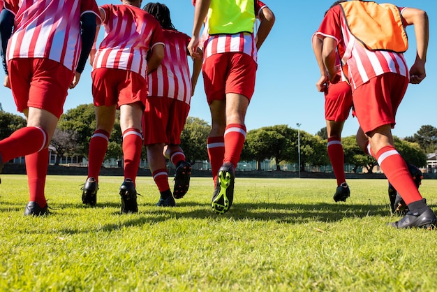 Vista baja de atletas masculinos multirraciales con uniformes rojos y zapatos de fútbol corriendo en tierra herbácea