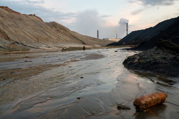 Vista de la carretera sucia con barro y grandes charcos o río rodeado de colinas contaminadas y fábricas industriales que exhalan humos tóxicos