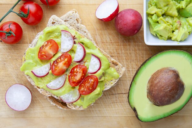 Vista cenital de pan tostado con aguacate, tomate y rábano en mesa de madera