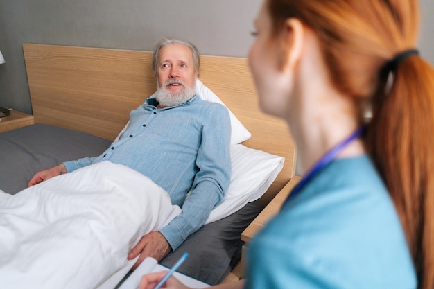 Vista de cerca desde atrás de una doctora sonriente hablando con un paciente masculino mayor acostado en la cama en el hogar de ancianos