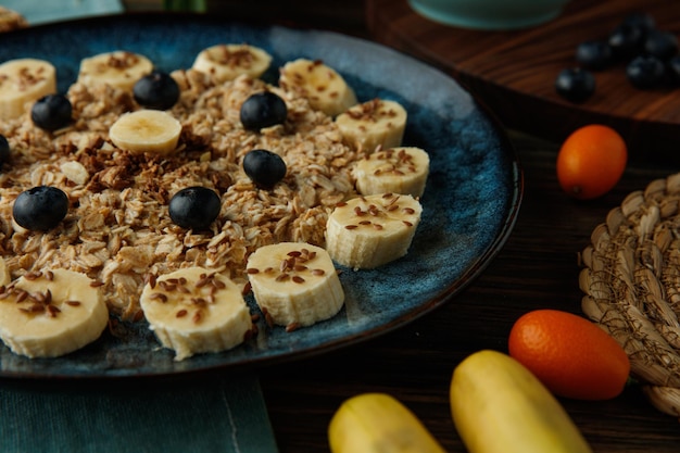 Vista de cerca de la avena con nuez de endrino de plátano y sésamo en un plato con plátano kumquat sobre tela azul sobre fondo de madera