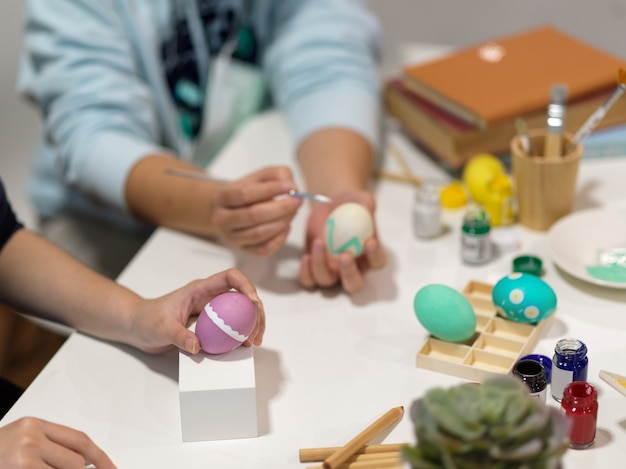 Vista de cerca de dos personas manos pintando huevos de Pascua con herramientas de pintura sobre la mesa