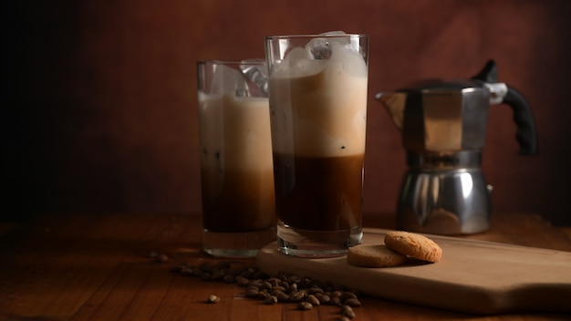 Vista de cerca de dos vasos de café helado con galletas en bandeja de madera, cafetera y granos de café decorados en la mesa