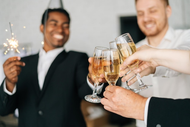 Foto vista de cerca del equipo de negocios de empleados multinacionales que sostienen luces de bengala, tintinean copas de champán y sonríen alegremente en año nuevo corporativo o feliz nochebuena.