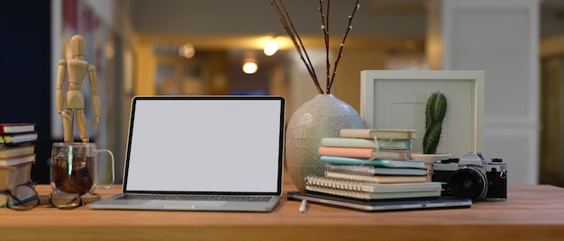 Vista de cerca del espacio de trabajo con computadora portátil de pantalla en blanco, libros, suministros y decoraciones en el escritorio de madera en la sala de estar