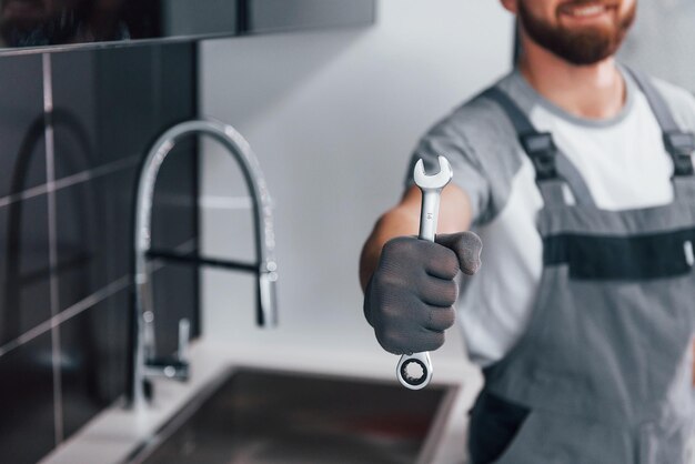 Vista de cerca de un joven plomero profesional con uniforme gris sosteniendo una llave en la mano en la cocina