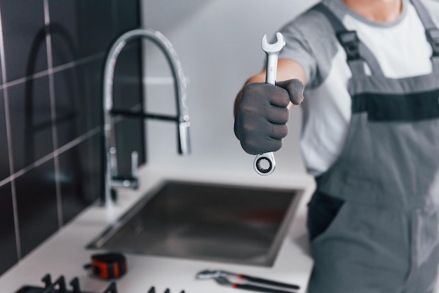 Vista de cerca de un joven plomero profesional con uniforme gris sosteniendo una llave en la mano en la cocina
