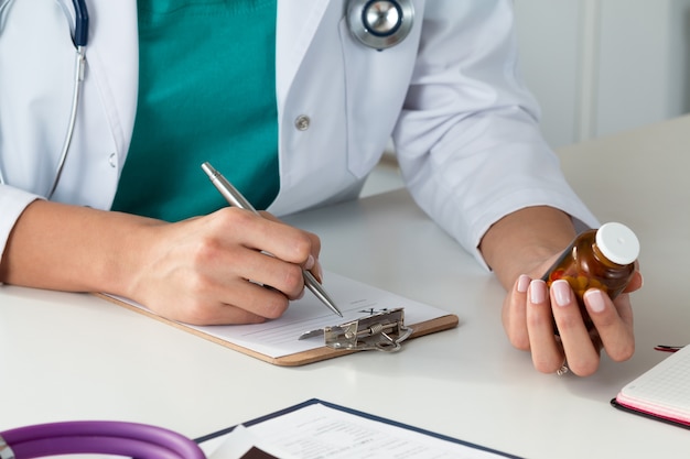 Vista de cerca de la mano de la doctora sosteniendo la botella con pastillas y prescripción de escritura