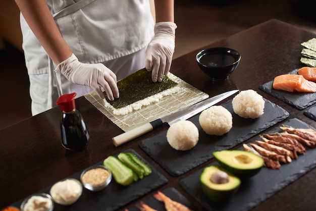 Vista de cerca de las manos de los chefs preparando sushi rodante con ingredientes arroz, nori, aguacate, salmón, salsa de soja