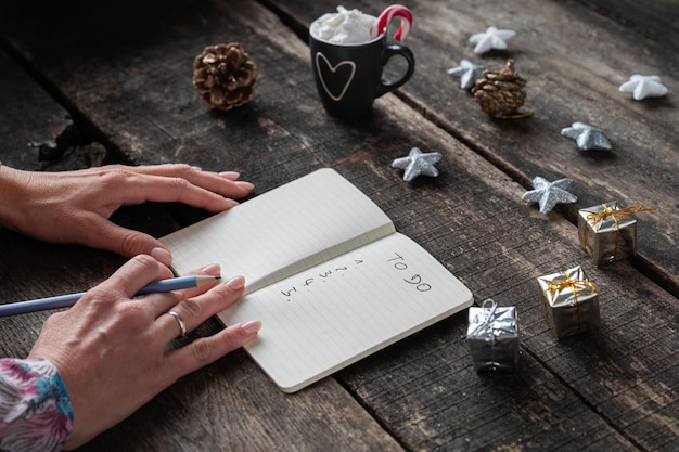 Vista de cerca de las manos femeninas escribiendo una lista de tareas pendientes en un bloc de notas en un escritorio de madera decorado con adornos navideños.